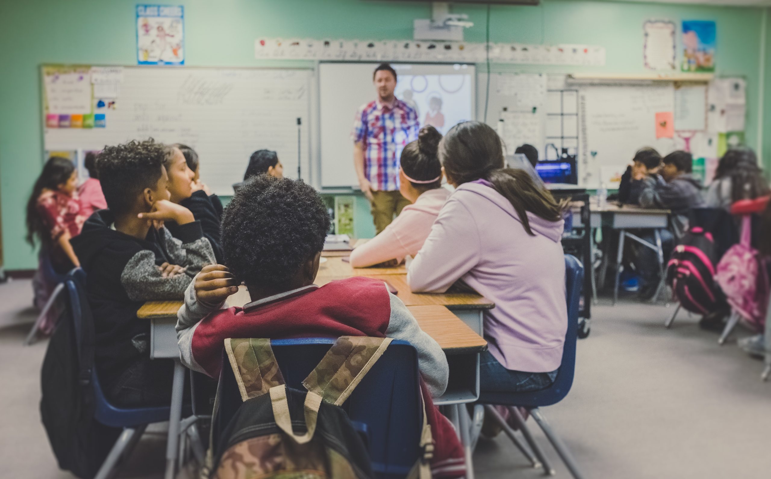 Students in a classroom listening to a teacher at the front of the room.